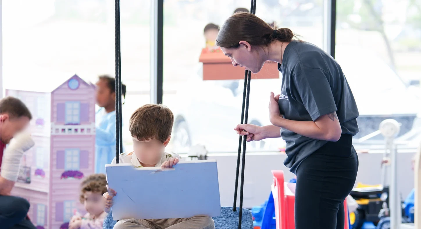 A Novinway Care Specialist leaning in to talk with a boy sitting on a platform swing